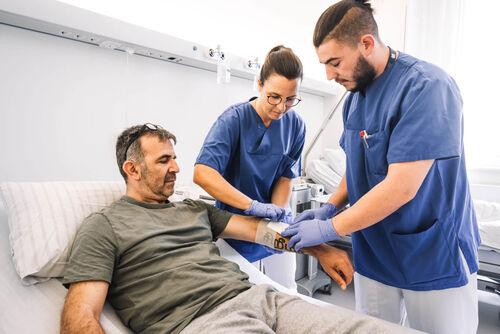 Das Bild zeigt eine pflegerische Mitarbeiterin und eine männliche Pflegefachkraft bei der Behandlung eines im Bett liegenden Patienten.