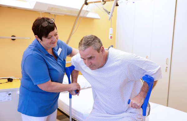 Eine Physiotherapeutin hilf einem Patienten beim Aufstehen aus dem Bett.